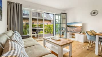 A modern living room with a sofa, a coffee table, and a TV. There is a balcony with a view of a brick building and greenery.