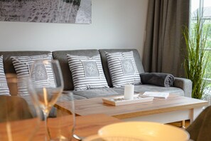 A modern living room with a grey sofa, striped pillows, a wooden coffee table, and a large window with curtains. - Beachfront accommodation for 5 people in a new building with a terrace and private sauna - (Hörnum (Sylt))
