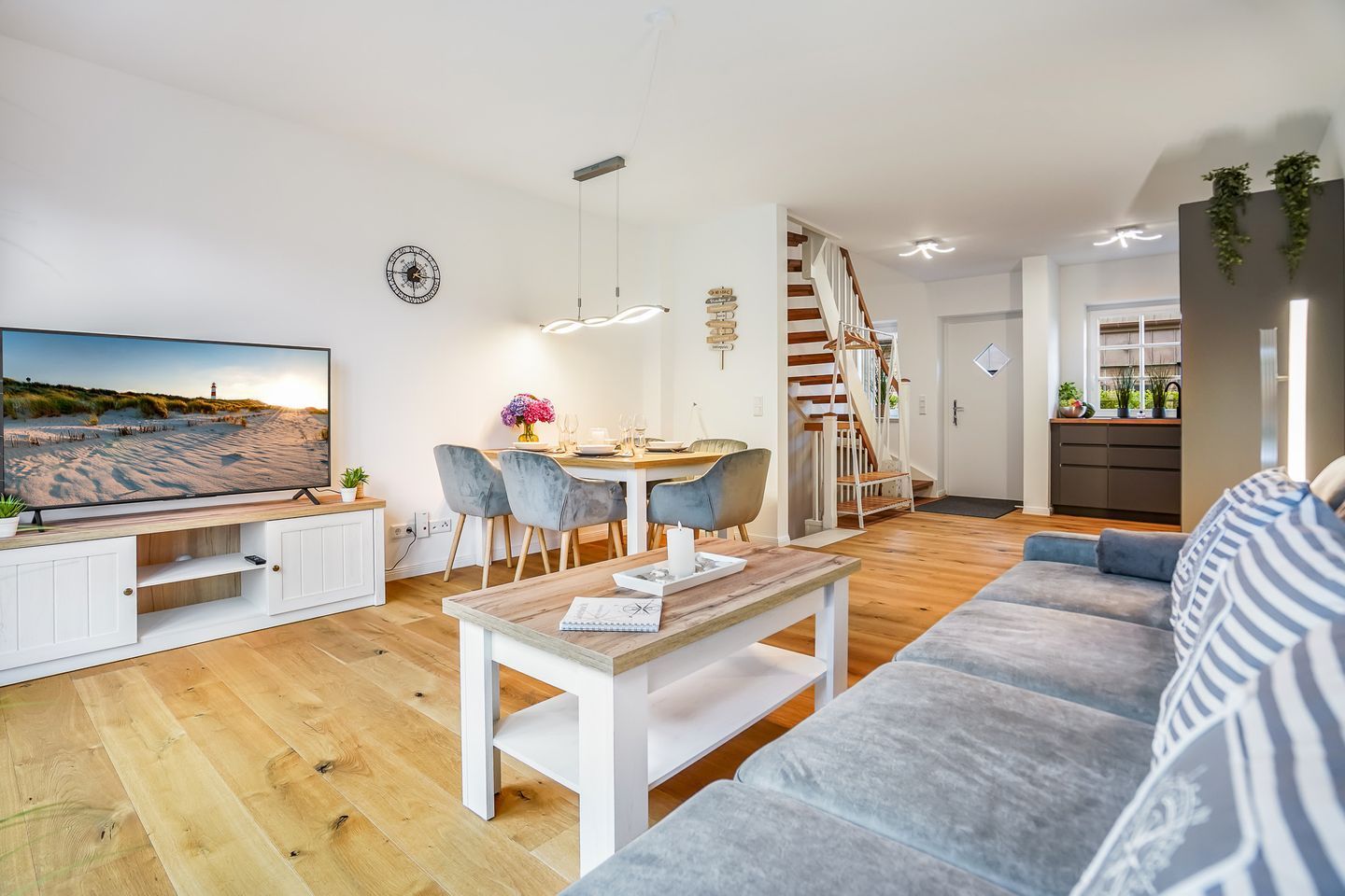 A modern living room with a flat-screen TV, a dining area with a wooden table and chairs, a staircase, and a kitchen area.