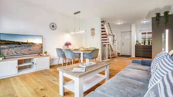 A modern living room with a flat-screen TV, a dining area with a wooden table and chairs, a staircase, and a kitchen area.