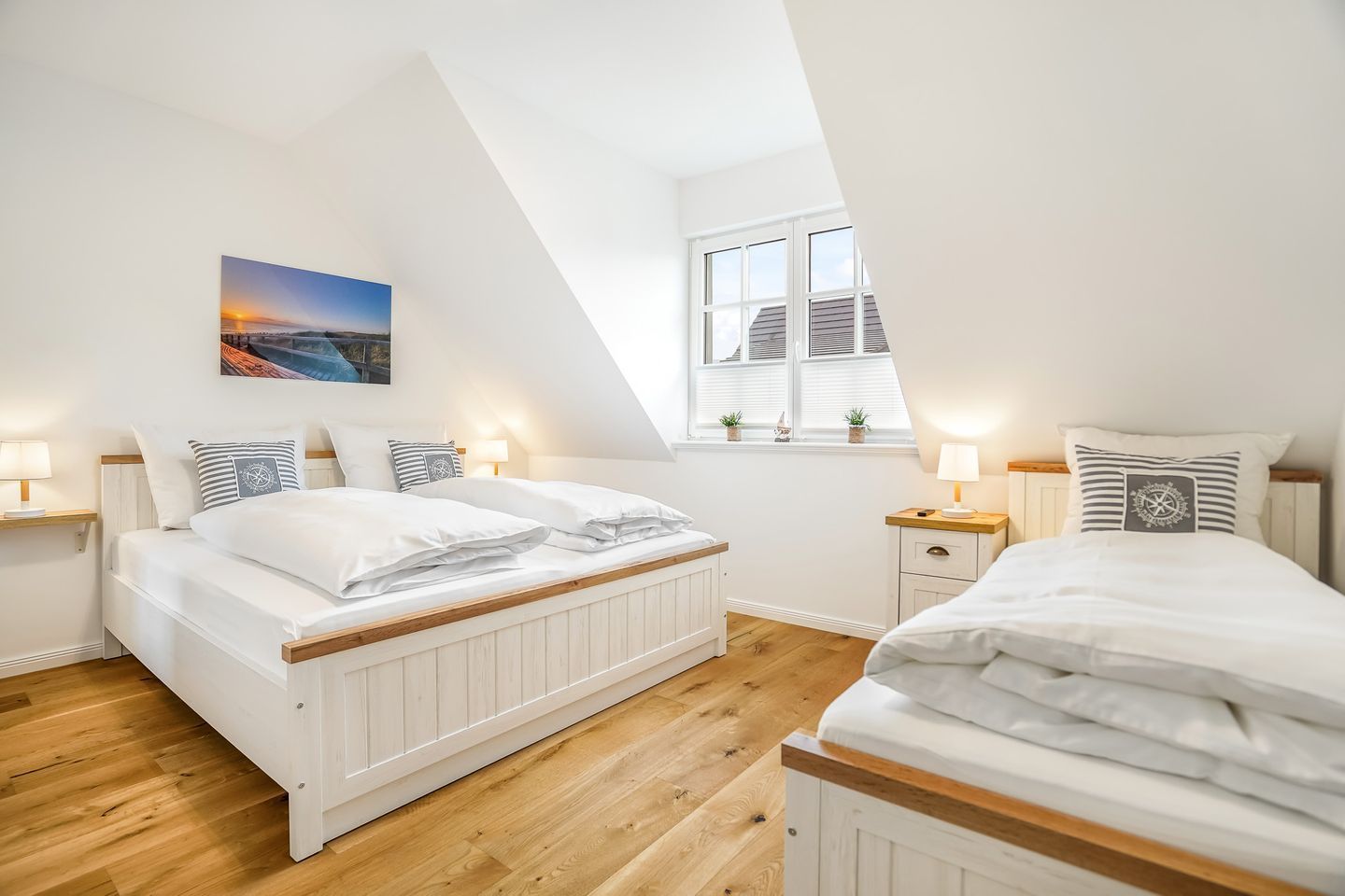 A bedroom with a sloped ceiling, a large bed with white bedding, two bedside tables with lamps, a window with blinds, and a painting on the wall.