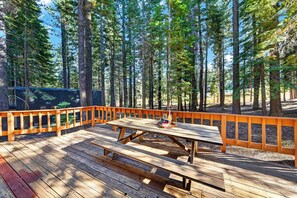 En träveranda med picknickbord och orangea räcken, omgiven av höga träd.