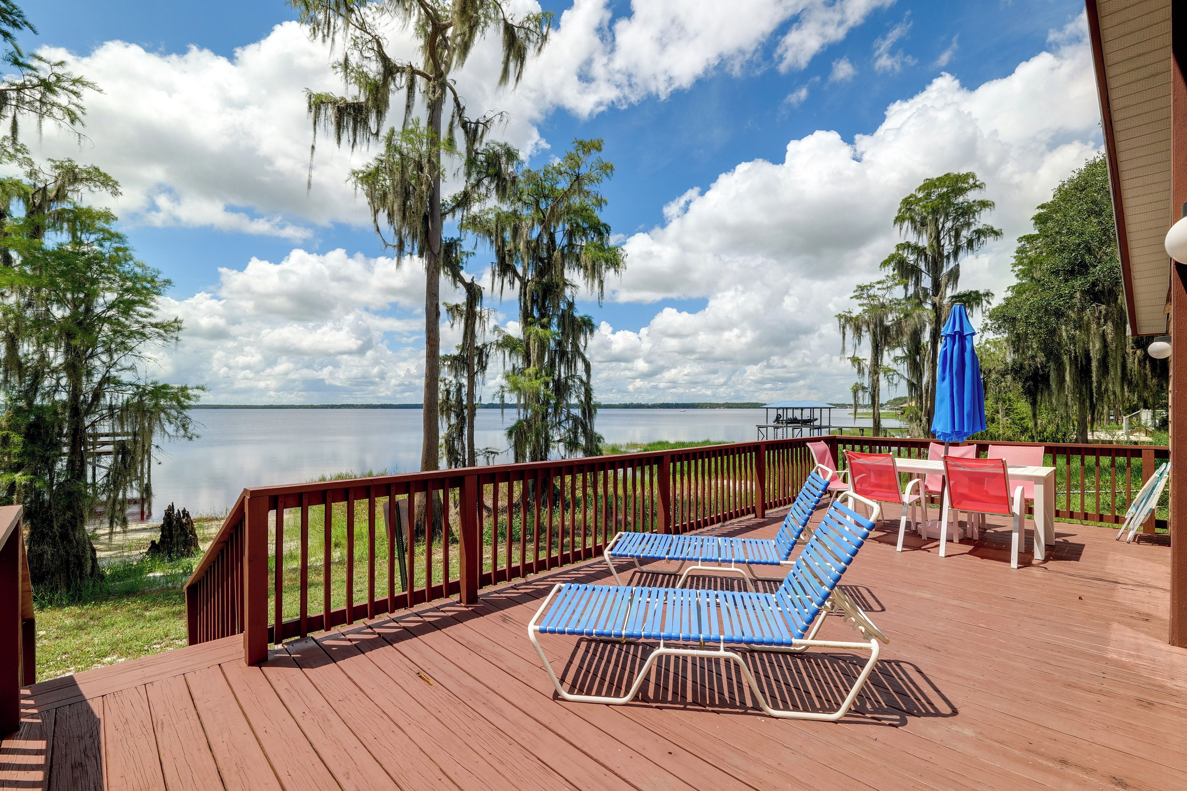 Une terrasse en bois avec des chaises longues bleues et une table, donnant sur un lac paisible bordé d’arbres et sous un ciel dégagé.