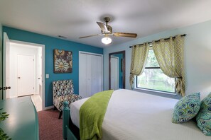 A bedroom with a bed, a chair, a ceiling fan, a window with curtains, and a door.