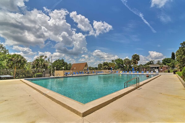 A rectangular swimming pool with clear blue water, surrounded by a concrete deck. There are lounge chairs aligned along the pool's edge, and a building with a brown roof is visible in the background.