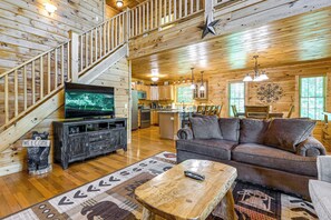 A wooden cabin interior with a staircase, a living room with a sofa and coffee table, a kitchen area, and a television. - 5 Mi to Cataloochee Ski! Mtn-View Retreat w/ Deck (Maggie Valley)
