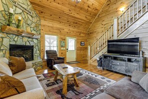 A cozy cabin interior with a stone fireplace, wooden walls, a comfortable sofa, a television, and a staircase. - 5 Mi to Cataloochee Ski! Mtn-View Retreat w/ Deck (Maggie Valley)