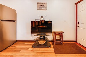 A room with a flat-screen TV on a stand, a wooden floor, a small wooden table, a wicker basket, and a door with a white frame. - Cozy Apartment Near UAF & Downtown Fairbanks! (Fairbanks)
