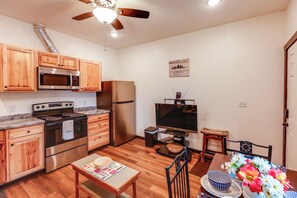 A compact kitchen with wooden cabinets, a stainless steel refrigerator, a microwave, and a stove. There's a dining table with a placemat and a bowl. A ceiling fan is visible above. - Cozy Apartment Near UAF & Downtown Fairbanks! (Fairbanks)