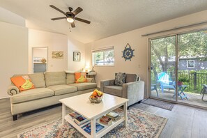 Ein Wohnzimmer mit einer Couch, einem Couchtisch mit Büchern, einem Deckenventilator und Blick durch eine Glastür auf einen Außenbereich mit Stühlen.