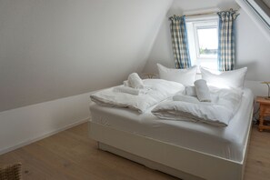 A neatly made bed in a room with a sloped ceiling and a window with checkered curtains. - Kapitänskoje - Comfortable Apartment in Keitum with a Beach Chair on the Beach at (Sylt)