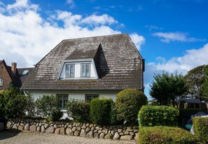 A two-story house with a sloped roof, a visible attic window, and a stone wall in the foreground. - Kapitänskoje - Comfortable Apartment in Keitum with a Beach Chair on the Beach at (Sylt)