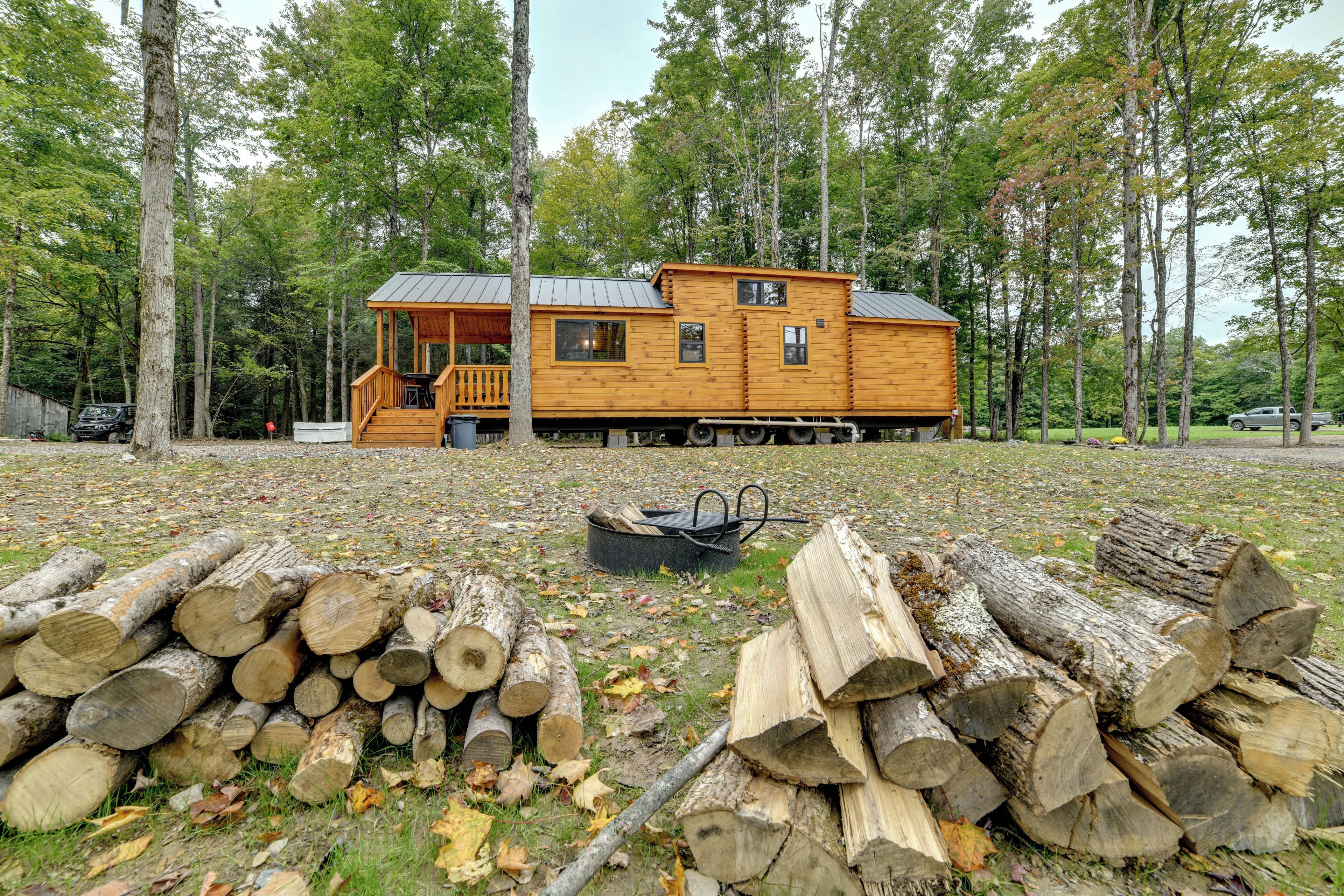 Une cabane en bois dotée d’un porche et d’une terrasse couverte, entourée d’arbres et d’une pile de bois de chauffage.