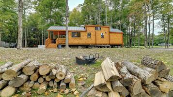 Une cabane en bois dotée d’un porche et d’une terrasse couverte, entourée d’arbres et d’une pile de bois de chauffage.