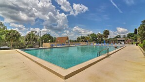 Et rektangulært swimmingpool med krystalklart blåt vand, omgivet af en betonflisebelagt terrasse. Der er liggestole langs poolens kant, og en bygning med et brunt tag kan ses i baggrunden.