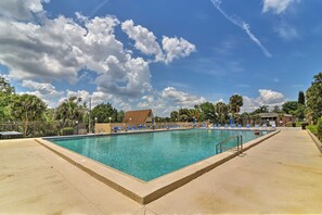 Une piscine rectangulaire à l’eau d’un bleu limpide, entourée d’une terrasse en béton. Des chaises longues sont alignées le long du bord de la piscine, et on aperçoit un bâtiment avec un toit brun à l’arrière-plan.