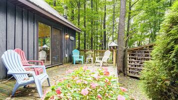 A wooden deck with Adirondack chairs, a glass door entrance, and a wooden fence with a stack of firewood.