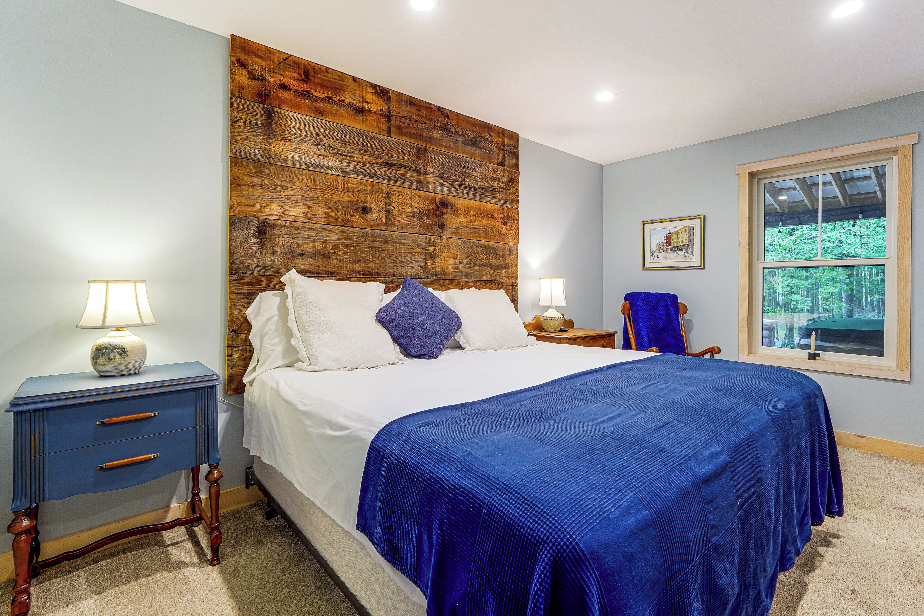 Une chambre avec une tête de lit en bois, un lit avec une literie blanche et un plaid bleu, une table de chevet avec une lampe, une commode avec des tiroirs et une fenêtre donnant sur des arbres.