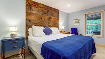 Une chambre avec une tête de lit en bois, un lit avec une literie blanche et un plaid bleu, une table de chevet avec une lampe, une commode avec des tiroirs et une fenêtre donnant sur des arbres.