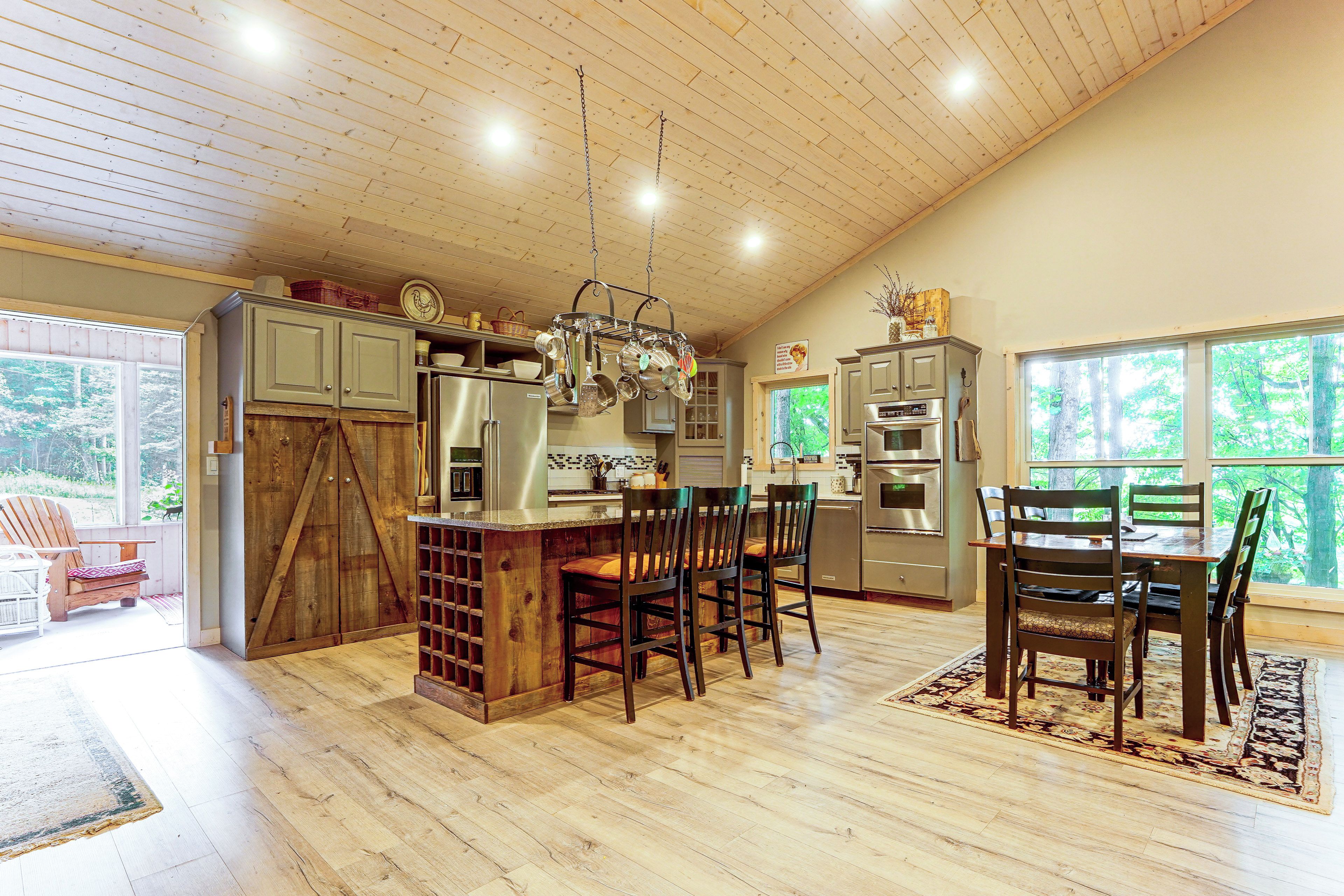A spacious kitchen with a wooden ceiling, a dining area with chairs, and a view of the outdoors through large windows.