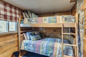 Una cama individual con colcha acolchada y almohadas, paredes de madera, una cortina a cuadros y una ventana con vistas a árboles.