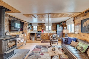 Interior acogedor de una cabaña con chimenea, muebles de madera, una zona de cocina y un ventanal que deja entrar mucha luz natural.