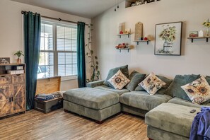 Ein Wohnzimmer mit einer Eckcouch, einer Holzkistenkommode, einem Fenster mit Vorhängen und Wandregalen mit Dekorationselementen.