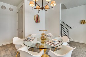 Un comedor con una mesa de vidrio rodeada de sillas blancas, una araña y una escalera al fondo.