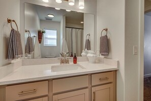 Un baño con un espejo grande, ganchos para toallas, un lavamanos con dispensador de jabón y un mueble con cajones.