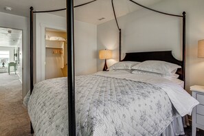 Ein Bett mit Baldachin, eine Bettdecke mit Blumenmuster, ein Nachttisch mit Lampe und ein begehbarer Kleiderschrank mit Kleidung.