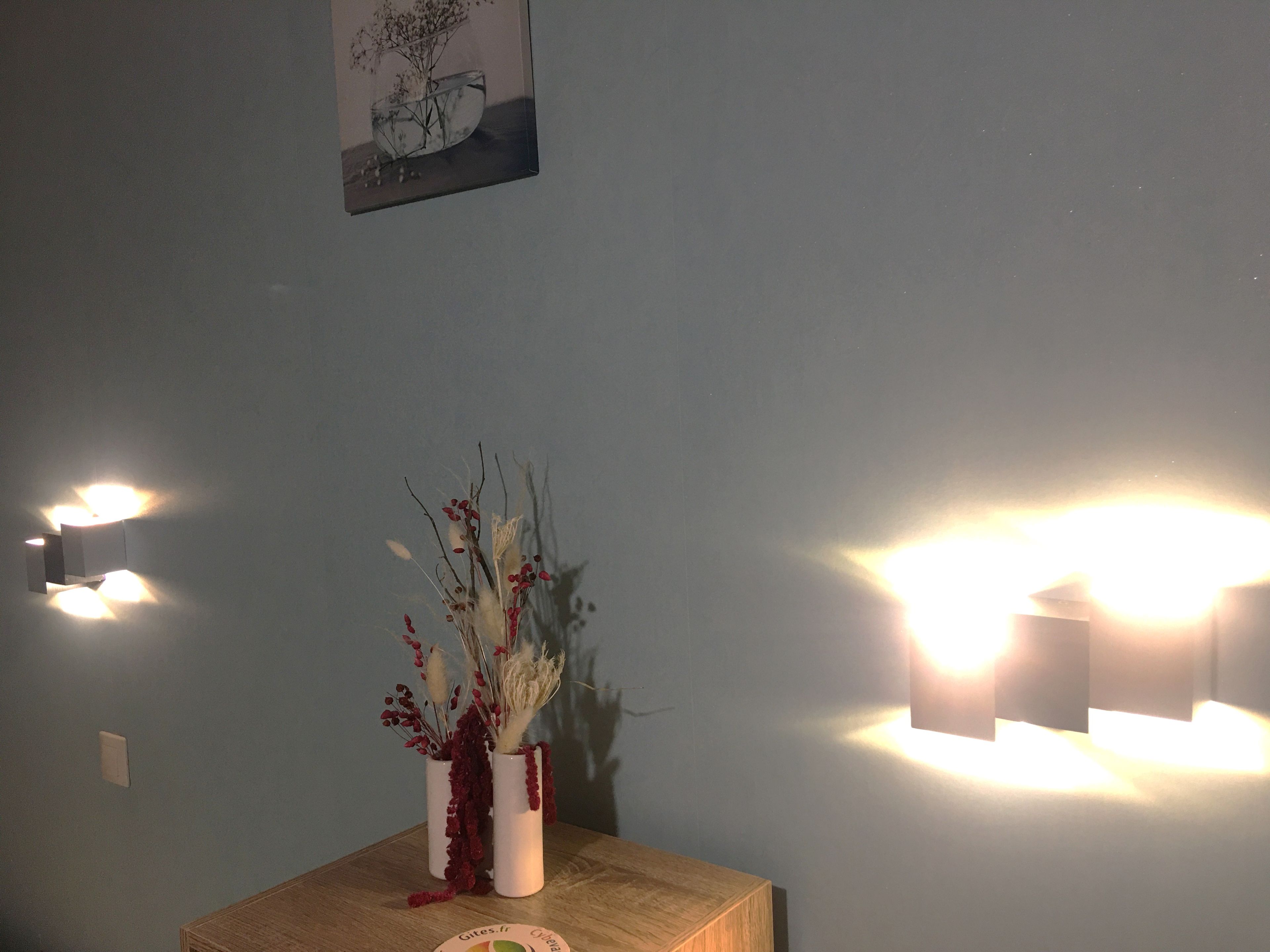 A room with a wooden table, a vase with dried flowers, and wall-mounted lights.