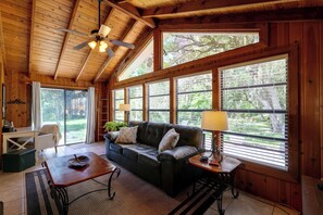 Ein gemütliches Zimmer mit Holzbalkendecke, einem Ledersofa, einem Holztisch und großen Fenstern mit Jalousien.
