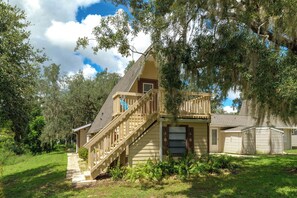 A two-story house with a wooden staircase leading to a balcony, surrounded by lush greenery and trees. - 'Good Vibes Cottage' Pool & Pier on Lake Louisa (Clermont)