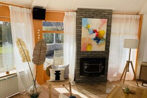 A living room with a fireplace, a large abstract painting, and a view of the outdoors. - Bungalow with 1 bedroom (Den Helder, Julianadorp)