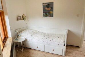 A small, single-bed room with a white bed, a bedside table, and a wall-mounted shelf with a doll and a picture frame. - Bungalow with 1 bedroom (Den Helder, Julianadorp)