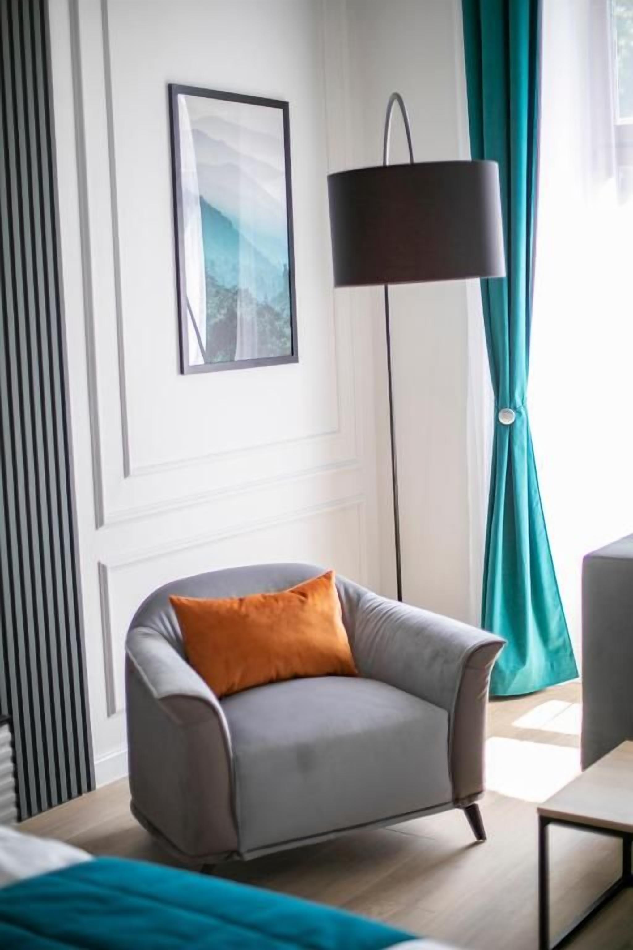 Una habitación moderna con un sillón gris, una lámpara de pie, un cuadro enmarcado en la pared y cortinas color esmeralda.