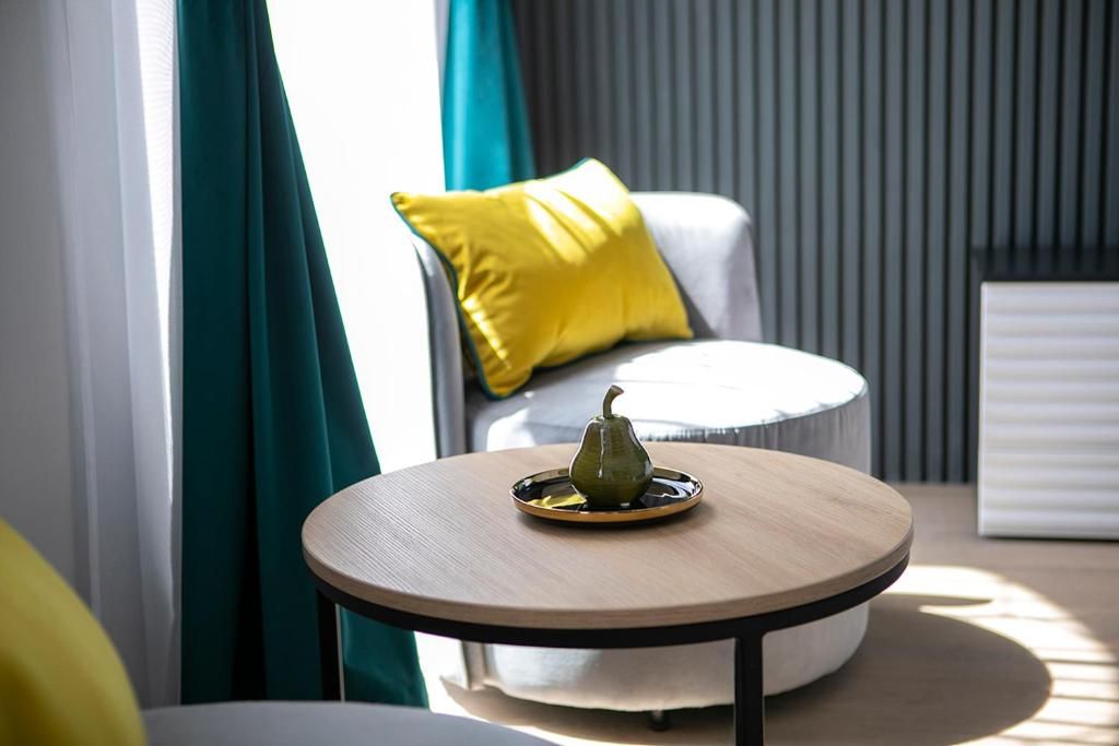 Una mesa redonda de madera con una pera verde sobre una bandeja, un sofá blanco con un cojín amarillo y cortinas color turquesa en una habitación moderna.