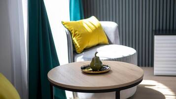 Una mesa redonda de madera con una pera verde sobre una bandeja, un sofá blanco con un cojín amarillo y cortinas color turquesa en una habitación moderna.
