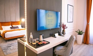 A modern hotel room with a bed, a desk with a flat-screen TV, a coffee maker, and a vase with flowers.