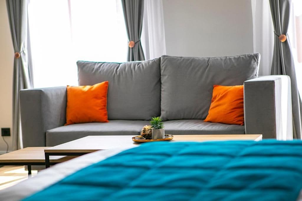 Una sala de estar moderna con un sofá gris, dos cojines naranjas, una planta pequeña sobre una bandeja y una manta turquesa en la cama.