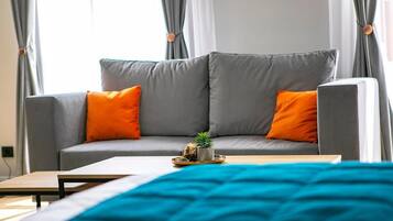 Una sala de estar moderna con un sofá gris, dos cojines naranjas, una planta pequeña sobre una bandeja y una manta turquesa en la cama.