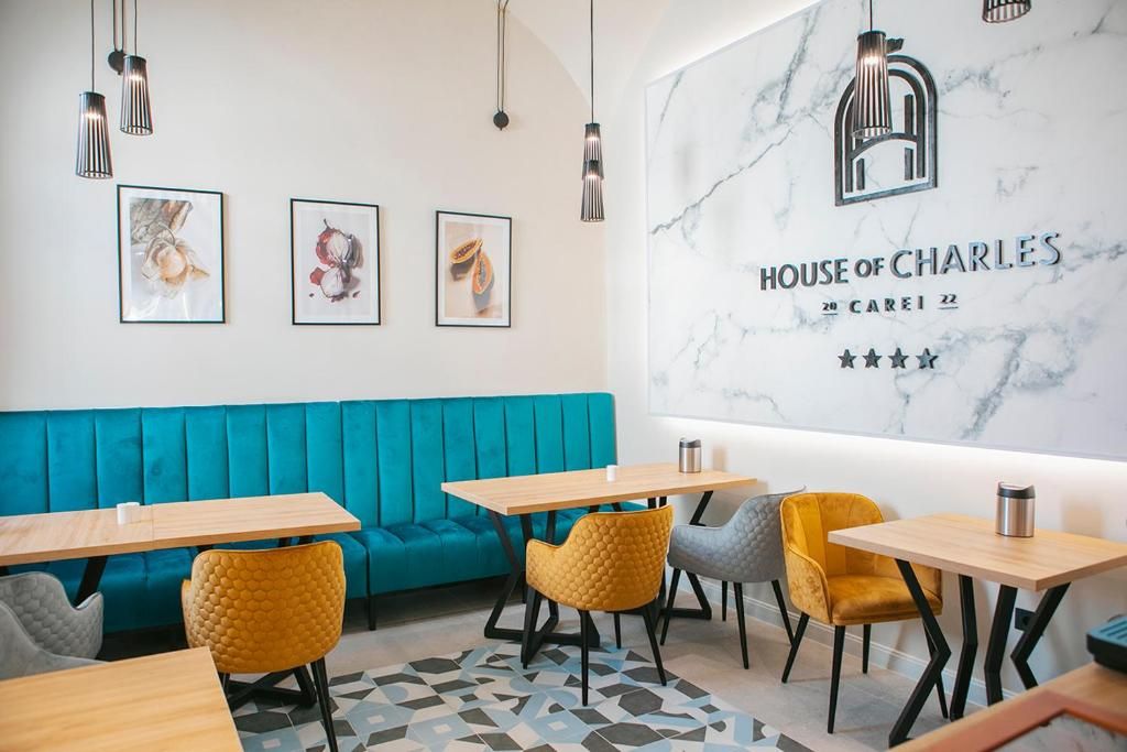 Un interior moderno con un sofá azul turquesa, mesas de madera y sillas amarillas. Se observa una pared de mármol con la señalización 'HOUSE OF CHARLES' y tres obras de arte enmarcadas.