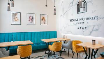 Un interior moderno con un sofá azul turquesa, mesas de madera y sillas amarillas. Se observa una pared de mármol con la señalización 'HOUSE OF CHARLES' y tres obras de arte enmarcadas.