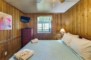 Ein Zimmer mit Holzwänden, einem Bett mit weißen Leinentüchern, einem an der Wand montierten Fernseher, einem Fenster mit Jalousien und einer Nachttischlampe.