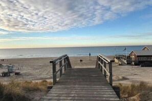 Ein hölzerner Steg führt zu einem Strand, auf der rechten Seite befindet sich ein Gebäude.
