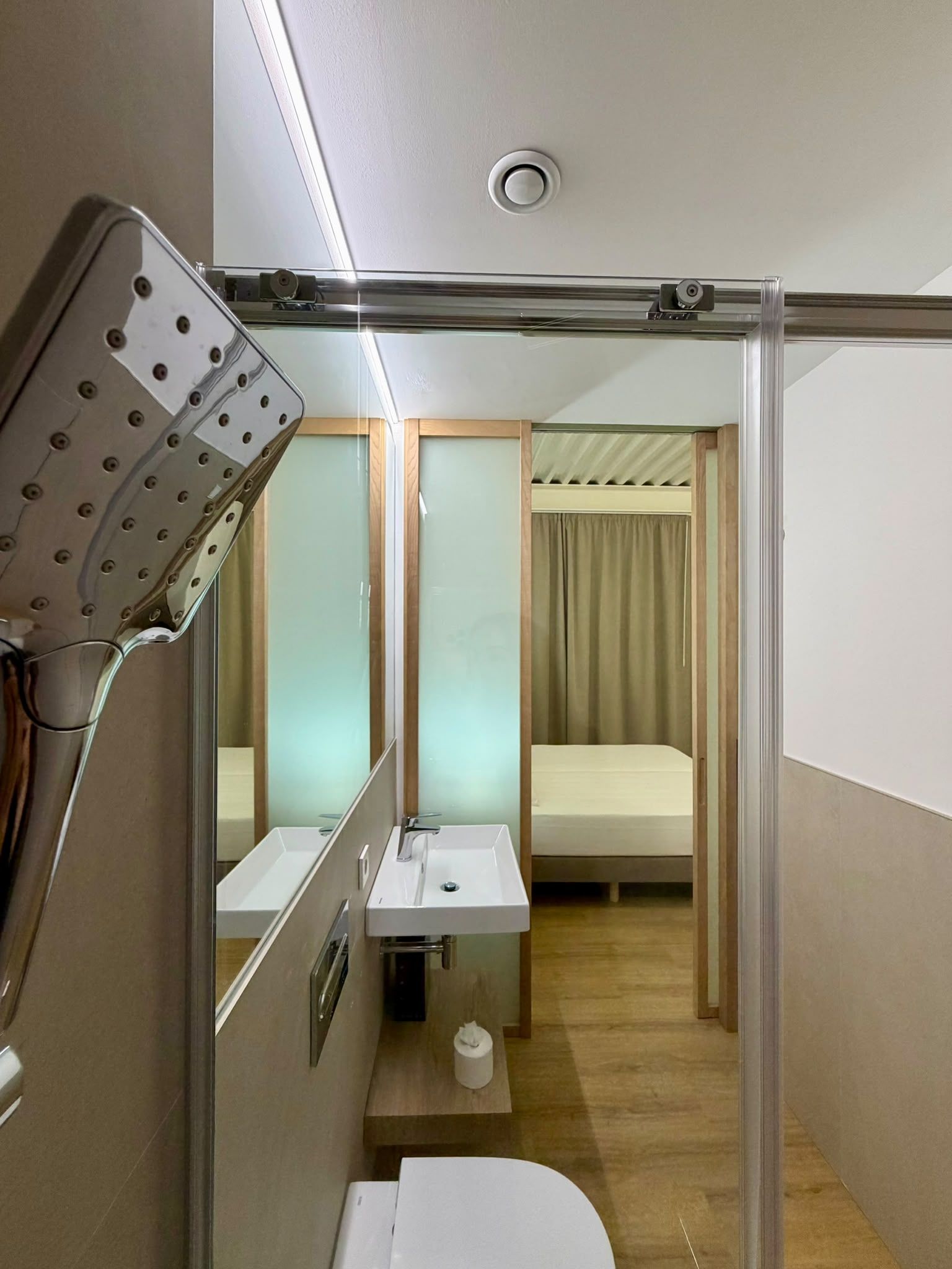 A compact hotel room with a shower, a small sink, a toilet, and a bed.