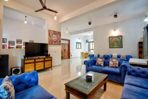 A spacious living room with a large flat-screen TV, blue sofas, a glass coffee table, and a ceiling fan. - Lux Beachfront 2 BHK Private Pool Villa in Morjim | 100mtr to Beach (Morjim)
