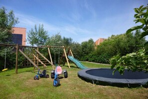 Een speeltuin met een schommel, glijbaan, driewieler en een trampo.