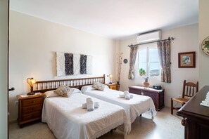 A hotel room with two beds, a wooden dresser, a desk, a chair, a window with curtains, a wall clock, and a wall-mounted air conditioner. - Villa in Nerja With Private Pool & Jacuzzi (Nerja)