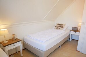 Ein Einzelbett mit weißer Matratze und einem Kissen mit Schriftzug, ein Nachttisch mit Lampe sowie ein kleiner Holfnachtschrank mit einem Behälter voller dekorativer Steine.